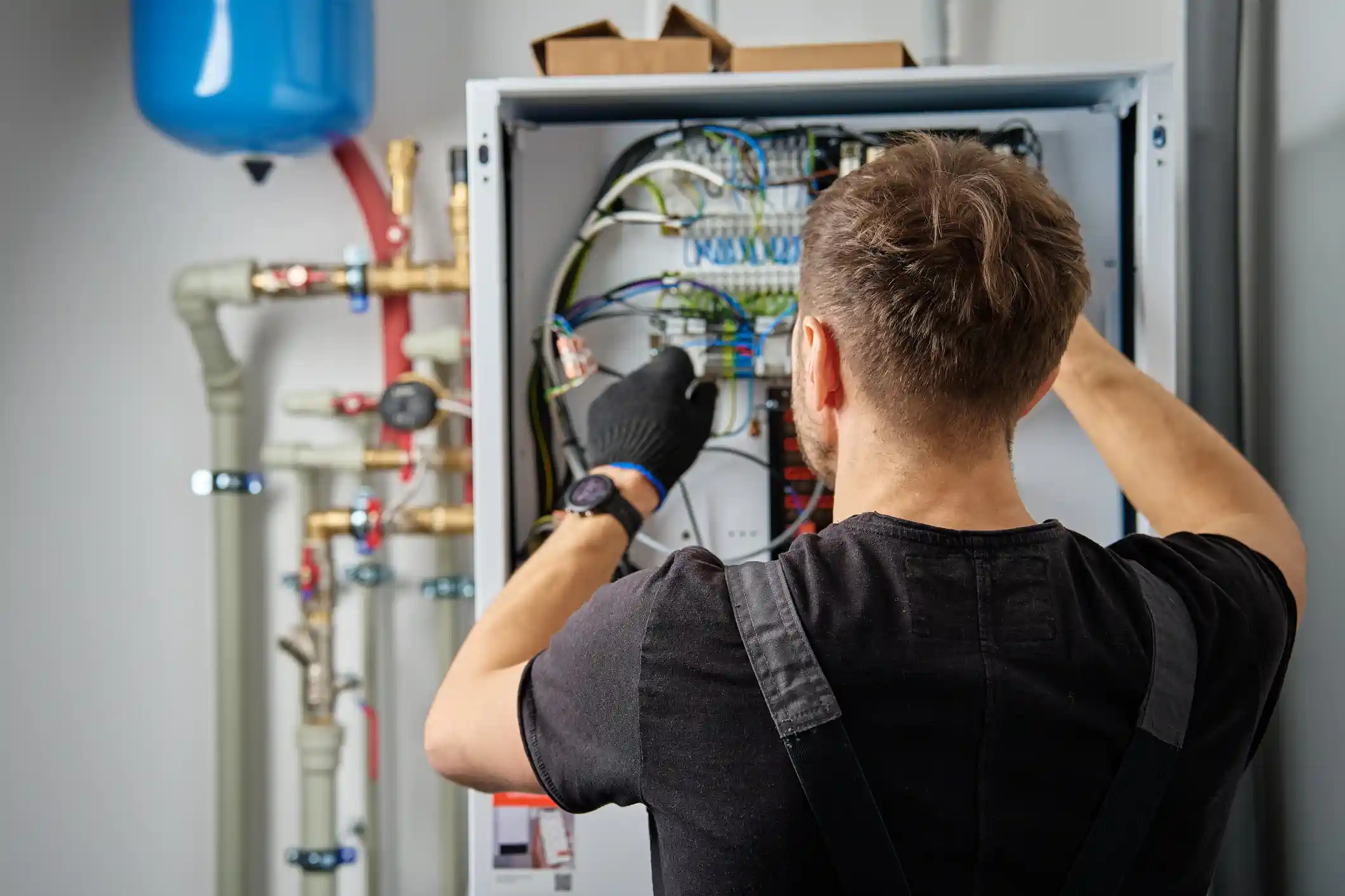 A rear-view close-up shot shows a male technician wearing a dark t-shirt, overalls, and a black work glove working on the internal electrical wiring of a large, wall-mounted white control box or indoor heat pump unit. He is reaching into the unit, connecting or adjusting various colored wires attached to a terminal block. In the blurred background to the left, there is a network of plumbing and pipes, including a prominent blue expansion tank, indicating the location is a utility room or boiler/mechanical closet. The image highlights electrical connection and maintenance for a heating system.