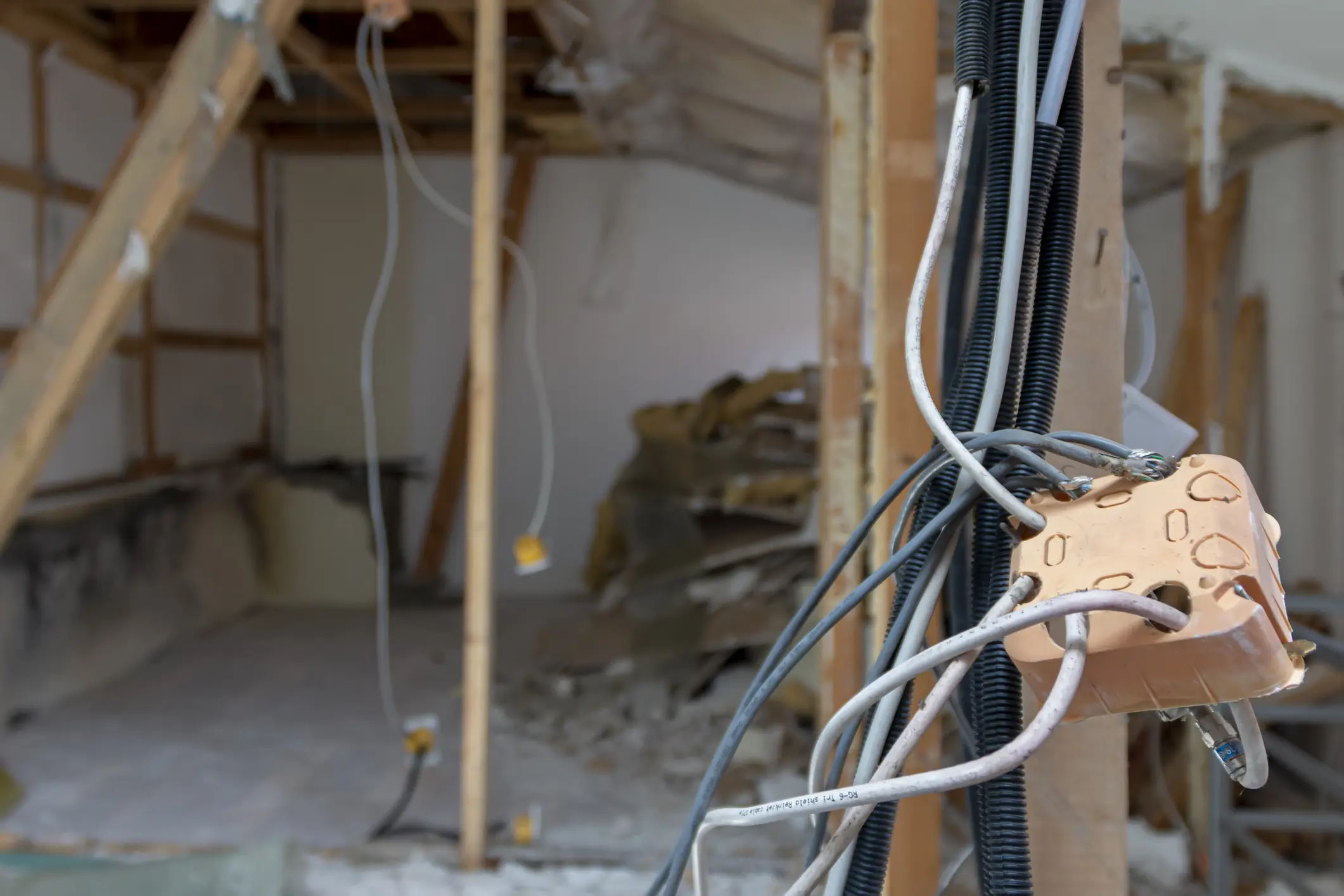 A close-up shot shows a tan-colored junction box overflowing with a mess of gray, black, and white electrical cables. The wires are being fed through black flexible conduit. In the blurred background, a partially demolished room is visible, with exposed wooden framing and construction debris on the floor, indicating a house rewiring project is underway.