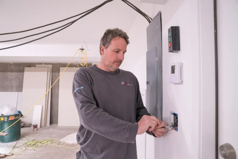  A man wearing a dark grey long-sleeved shirt with a logo on the sleeve (possibly "DAIKIN") is working on installing or connecting wiring to a wall outlet or switch in a room under construction. He has a light beard and is focused on the task.