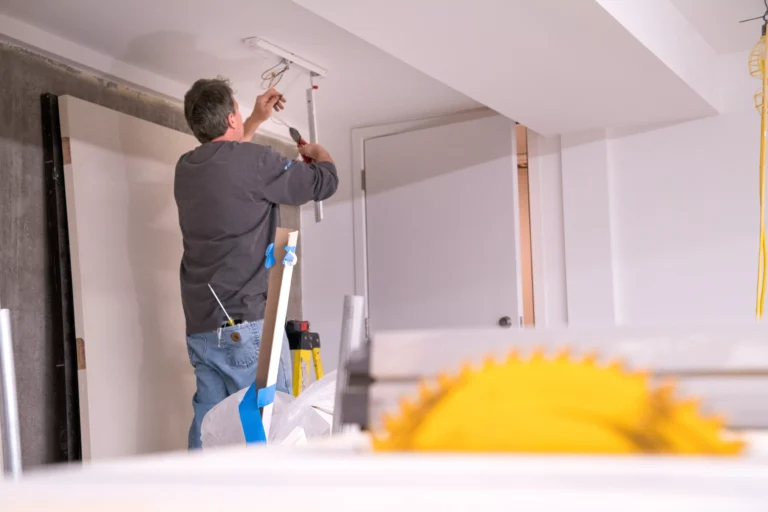 A man wearing a dark grey shirt and blue jeans is standing on a ladder, installing or working on a light fixture on a white ceiling in a room under construction. He is holding wiring and tools. In the background, there is a white door, white walls, and a large metal saw blade (yellow) is visible in the foreground, out of focus, suggesting a work site.