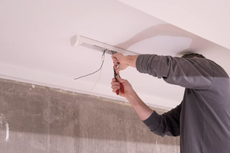  A man wearing a dark grey long-sleeved shirt is performing electrical work on a white light fixture mounted on a white ceiling. He is using red-handled wire cutters or strippers in his right hand to work with a bundle of black and white wires hanging from the fixture. Below the ceiling, the wall is unfinished, showing a rough, concrete or plaster texture.