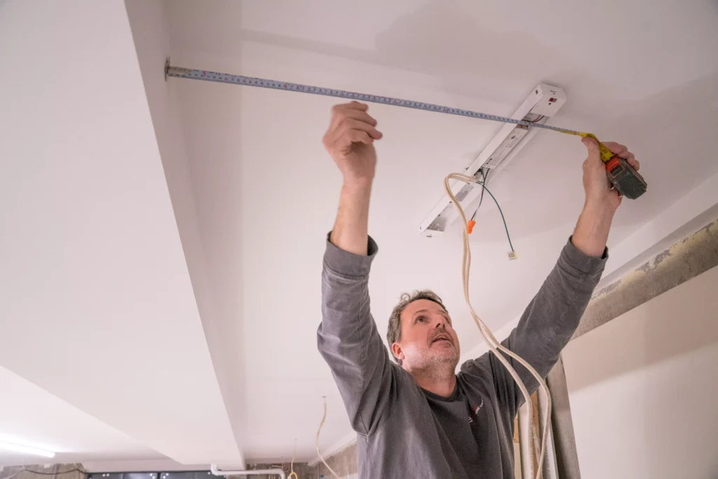 Electrician measuring for ceiling light installation in residential setting, showcasing electrical renovation services in Shakopee, MN.