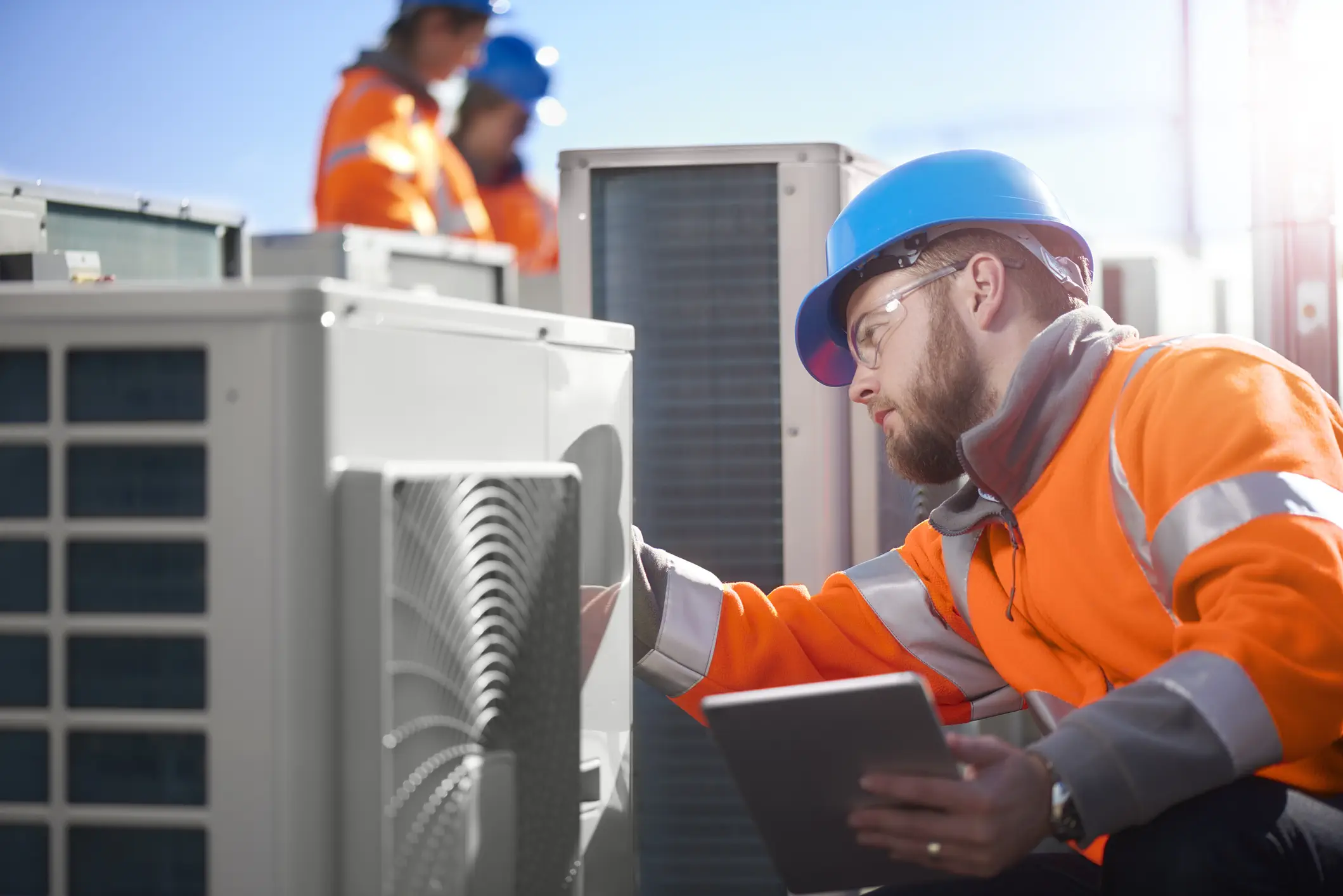 The image features an HVAC technician working on a bank of outdoor air conditioner condenser units or heat pumps on a rooftop, likely a commercial installation. The technician, with a beard and safety glasses, is wearing an orange high-visibility jacket and a blue hard hat. He is inspecting a unit and holding a digital tablet or clipboard, suggesting he is reviewing schematics or logging data. Two other technicians, also in similar safety gear, are visible in the blurred background. The scene is brightly lit by sunlight.