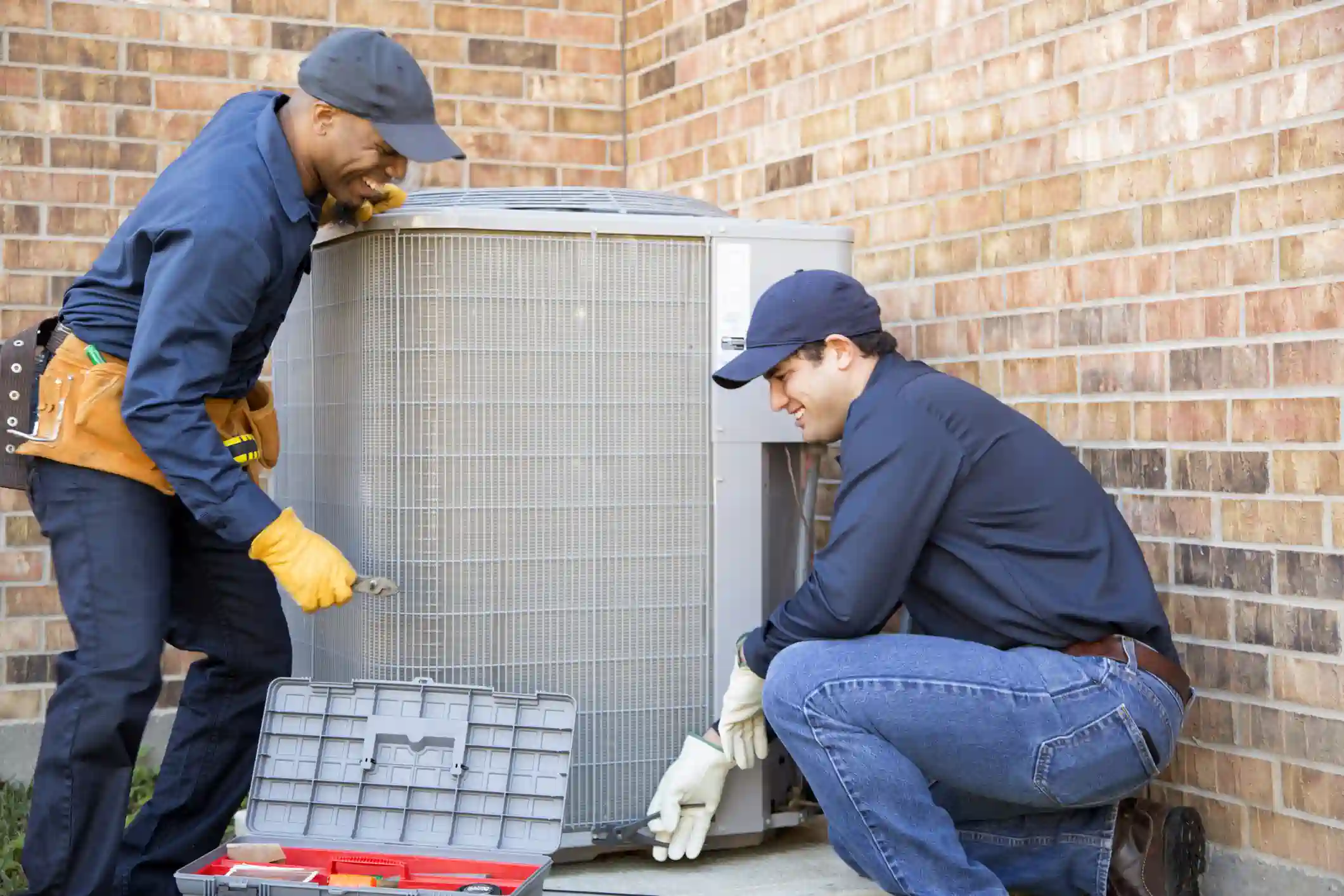 Two HVAC technicians are working on an outdoor air conditioning (AC) unit against a red brick wall. The technician on the left, who is Black, is standing, smiling, wearing a dark blue uniform, yellow gloves, and a tan tool belt, and holding a wrench. The technician on the right is crouched down, wearing a dark blue shirt, jeans, and light-colored gloves, and is also smiling as he works on the base of the unit. A gray toolbox is open in the foreground.