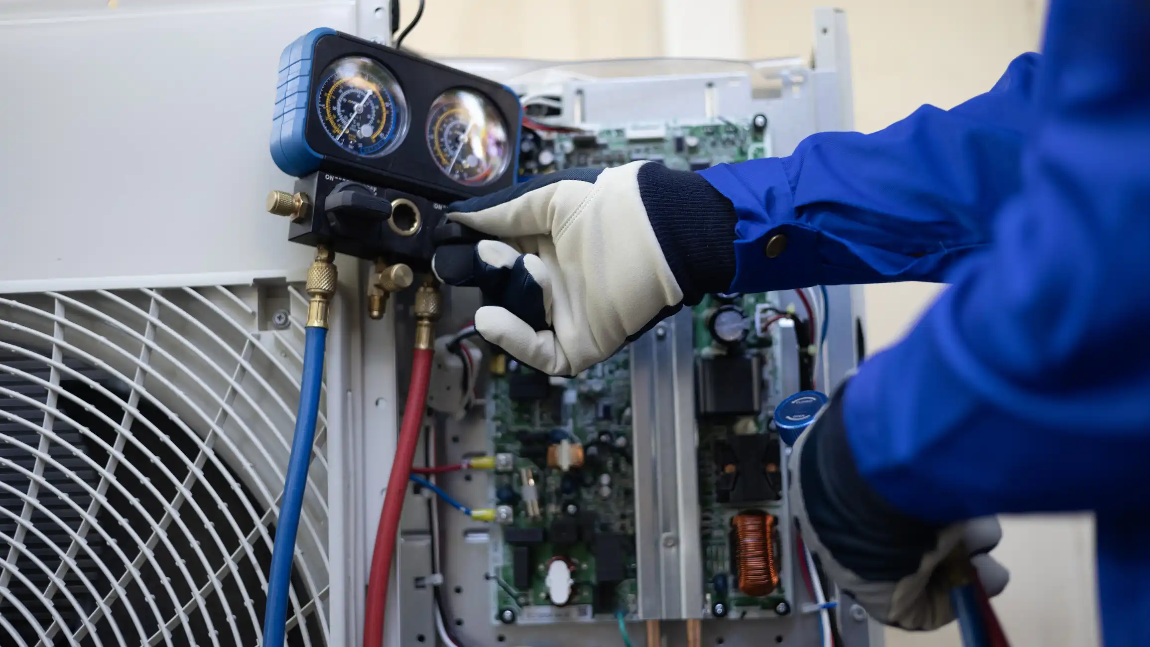  A close-up shot shows a technician, wearing a blue long-sleeved uniform and white work gloves, servicing the internal components of a white air conditioning (AC) unit. The technician is adjusting the valve on a refrigerant manifold gauge set (with blue and yellow gauges) which is connected to the unit's service ports by red and blue hoses. The unit's control board and electronics are visible where a panel has been removed, and the unit's fan guard is seen on the left.