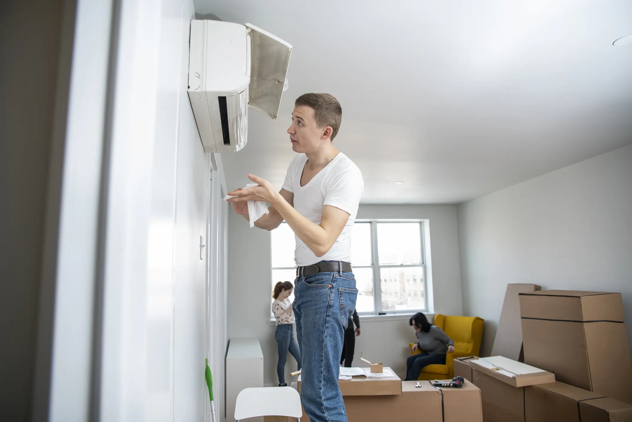 A young man in a white V-neck t-shirt and jeans is inspecting or servicing a white mini-split air conditioner unit mounted high on a white wall. The cover of the unit is open. He is wiping his hands with a cloth. The room appears to be an apartment with bare walls, large windows, and several cardboard moving boxes on the floor, suggesting a recent move or installation. Two people are visible in the background, one sitting on a yellow armchair.