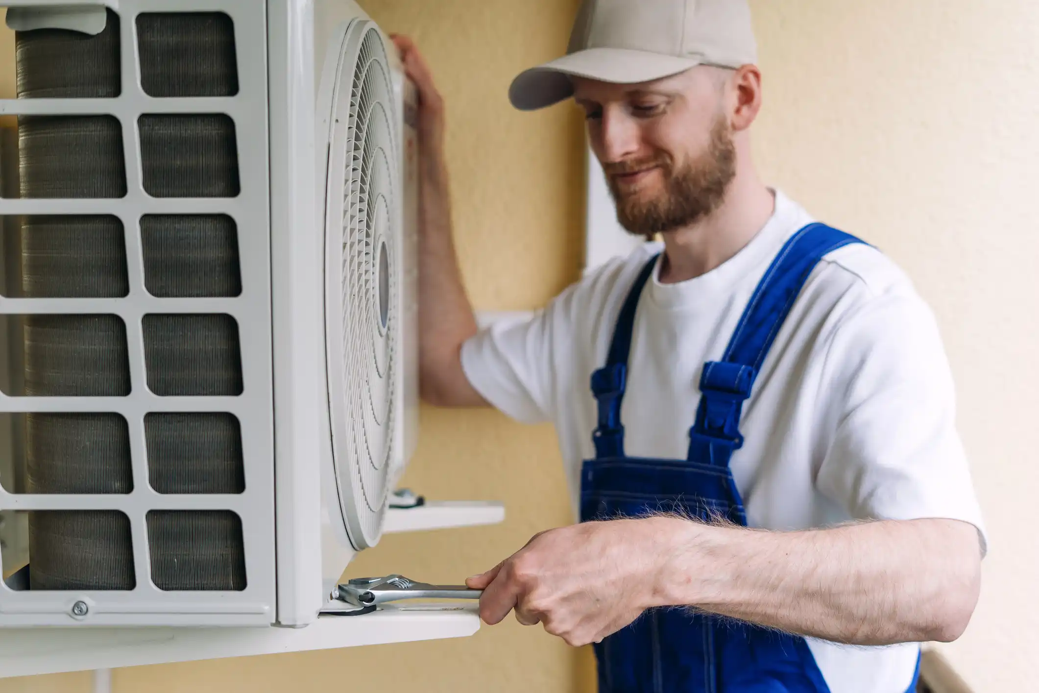 A smiling male technician with a beard and a beige baseball cap is servicing or installing an outdoor mini-split air conditioner unit. He is wearing a white t-shirt and blue overalls. He is using an adjustable wrench to tighten a fitting or bolt on the bottom right side of the unit, which is mounted on a light yellow wall.