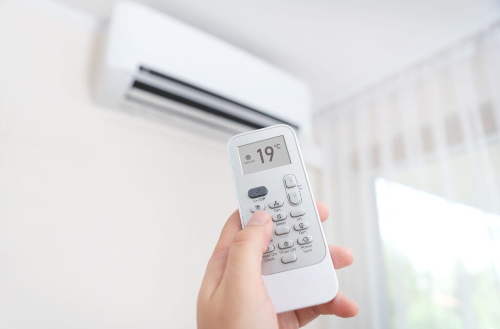 Hand holding air conditioner remote control displaying 19°C, with indoor unit mounted above, illustrating air conditioning service and comfort in Minneapolis, MN.