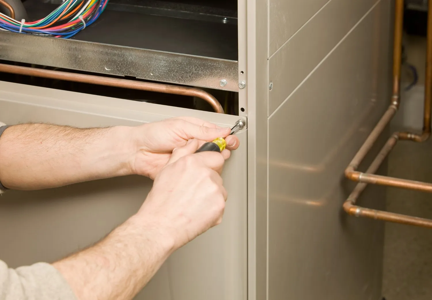 A close-up of a technician's hands using a yellow and black screwdriver to remove or fasten a screw on the beige metal access panel of a furnace