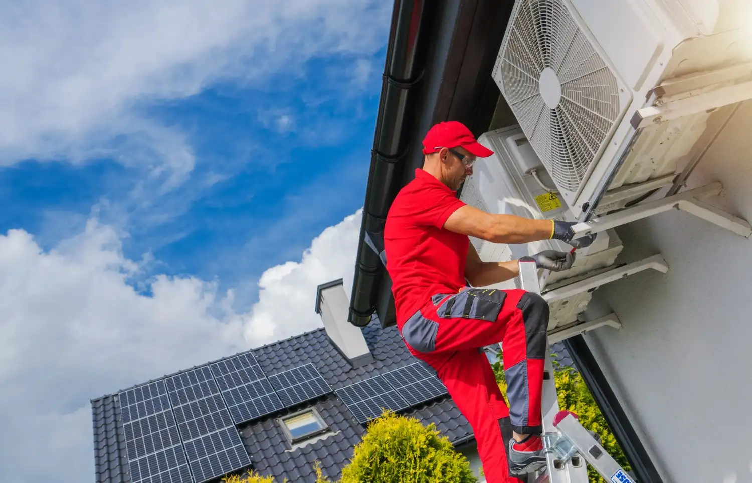 A technician in red workwear climbs a ladder to service a white air conditioning unit attached to a building under a partly cloudy sky