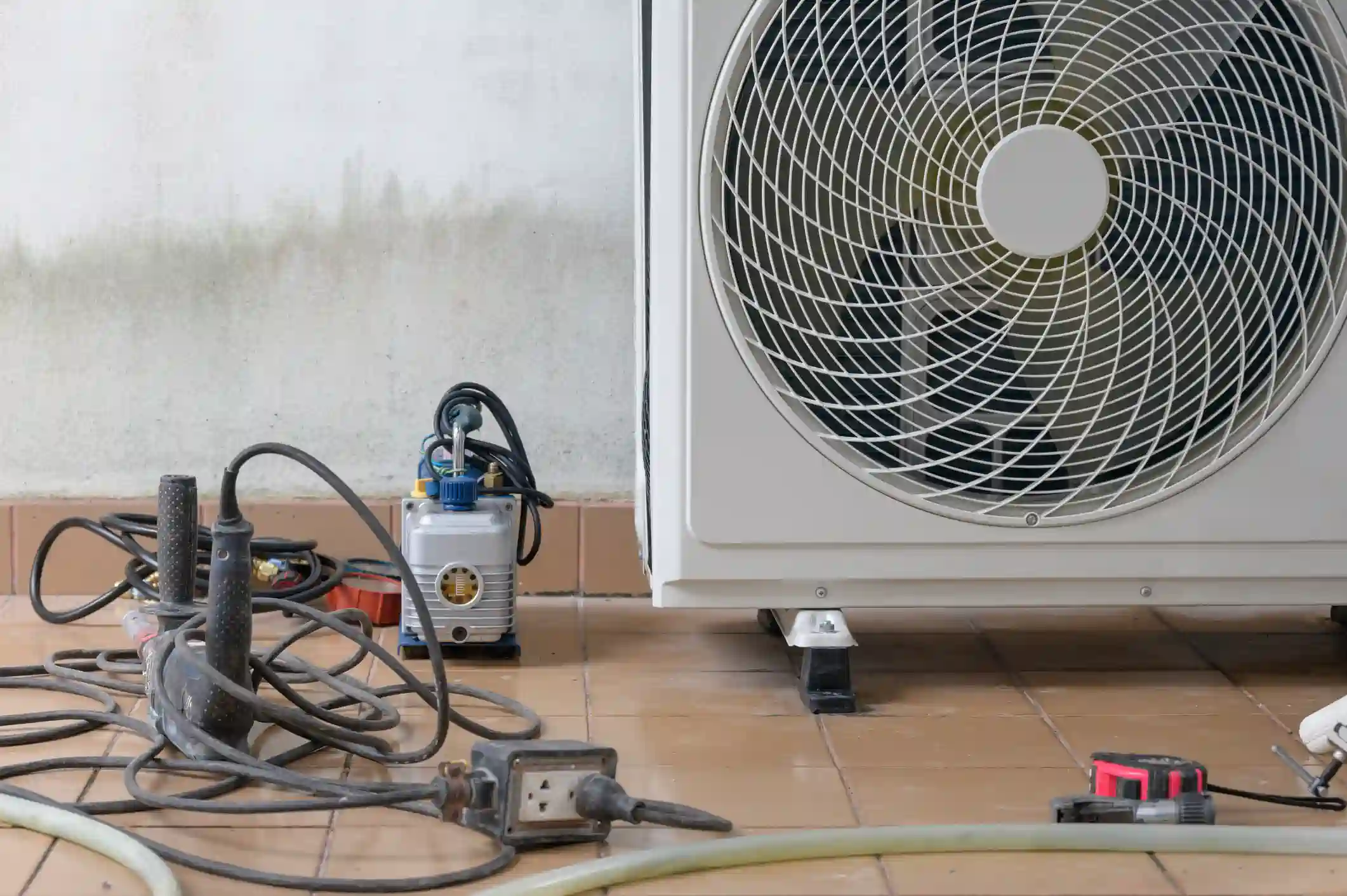 A low-angle close-up shot focuses on the bottom of an outdoor air conditioning or heat pump condenser unit and HVAC tools on a tiled patio floor. The white unit's fan grille and housing dominate the right side of the image. On the left, a grey vacuum pump, a tangle of black power cords and hoses, an extension cord plug, and a red and black measuring tape or tool are spread across the floor. The background is a grubby, light-colored wall. The image depicts the tools and equipment used for the installation or service of a refrigeration system.