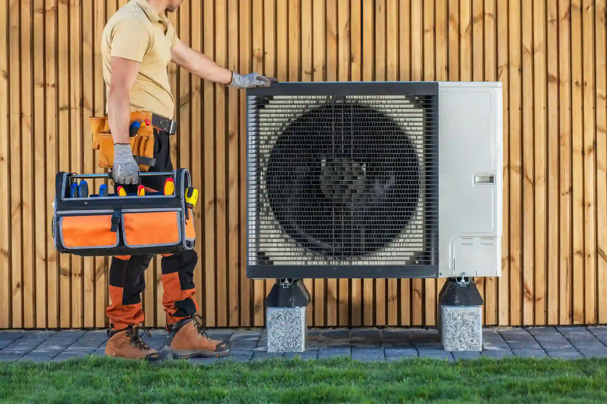  A professional image captures a male HVAC technician standing next to a large, modern air source heat pump unit. The technician wears a khaki polo shirt, two-toned orange and black work pants, work boots, a tool belt, and grey gloves. He is holding a black and orange soft-sided toolbox and touching the black protective grille of the heat pump. The unit is mounted on concrete blocks and set against a light-colored wooden slat wall. The foreground shows a grey brick patio and green grass. The scene suggests a professional arriving to perform heat pump service or maintenance.