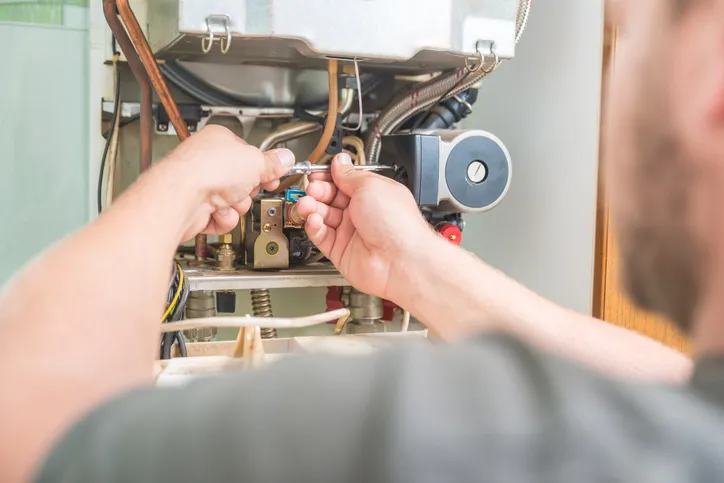 A person is using a screwdriver to adjust the internal components of a gas-fired boiler, with a focus on their hands and the intricate piping.
