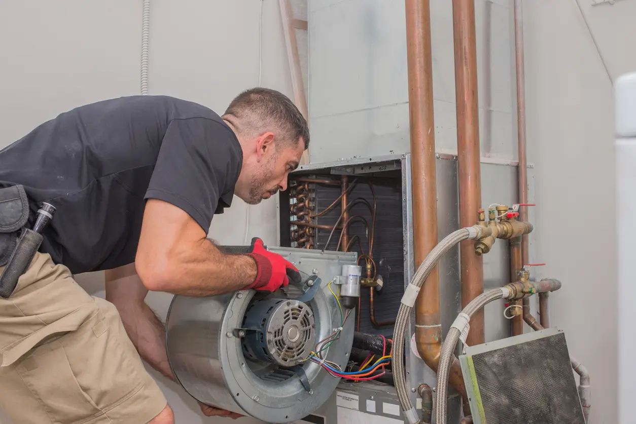 A maintenance technician, wearing a black shirt and khaki shorts, is bent over an indoor furnace unit. He is holding a blower motor and fan assembly, wearing a red and black work glove on his left hand. The evaporator coil and copper pipes are visible inside the opened unit.