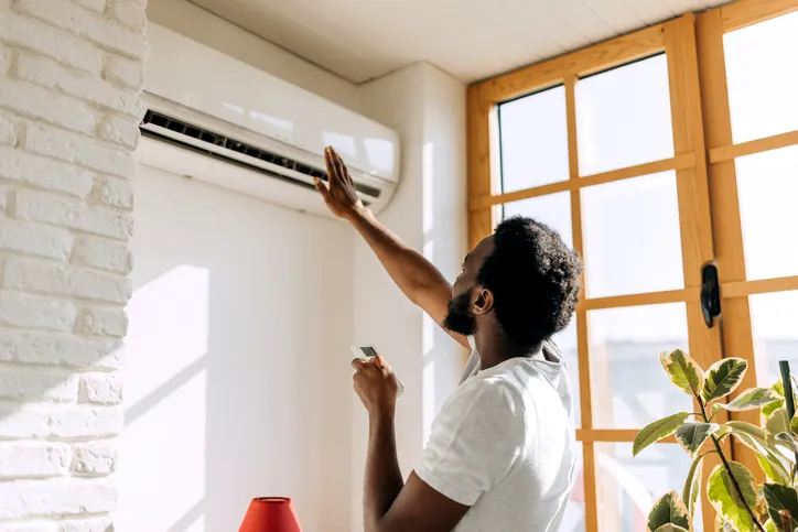 A person reaching up towards a wall-mounted mini-split air conditioner, holding a remote control