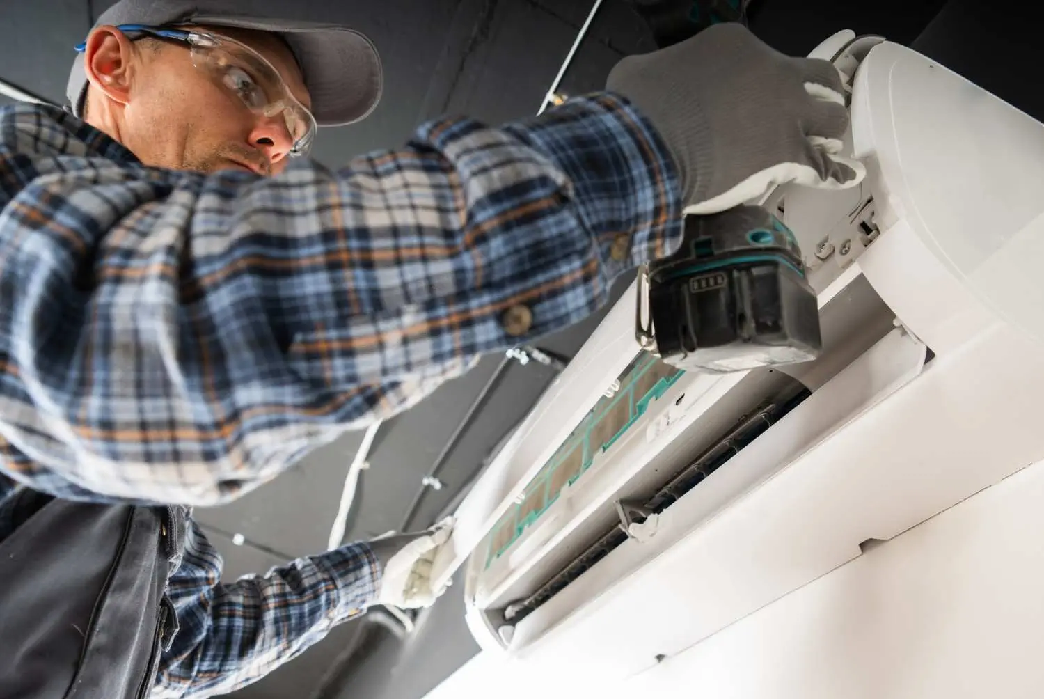 A low-angle close-up shot of an HVAC installer wearing a plaid shirt, gray cap, and safety glasses. He is wearing work gloves and is using a power drill to secure or disassemble the white casing of a ductless mini-split indoor unit mounted on a ceiling or high on a wall.