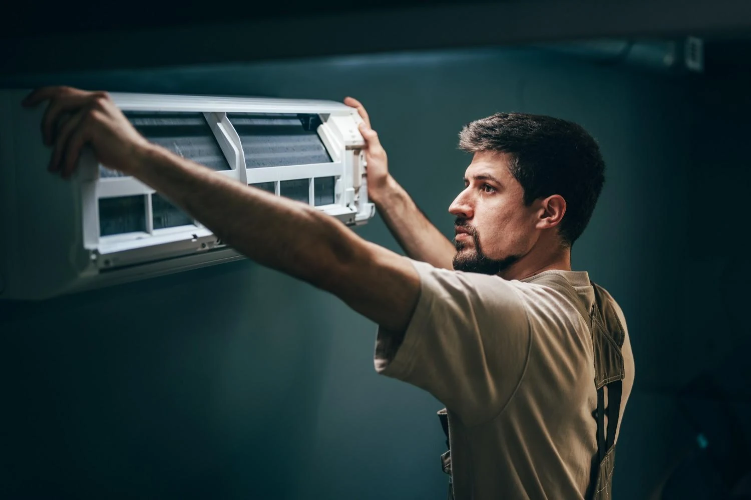 A bearded young man, an HVAC technician, wearing a light-colored t-shirt and overalls, is reaching up to service or install a ductless mini-split air conditioner indoor unit. He is holding the white casing open to access the internal components, with the air filters visible, against a dark teal or gray wall.