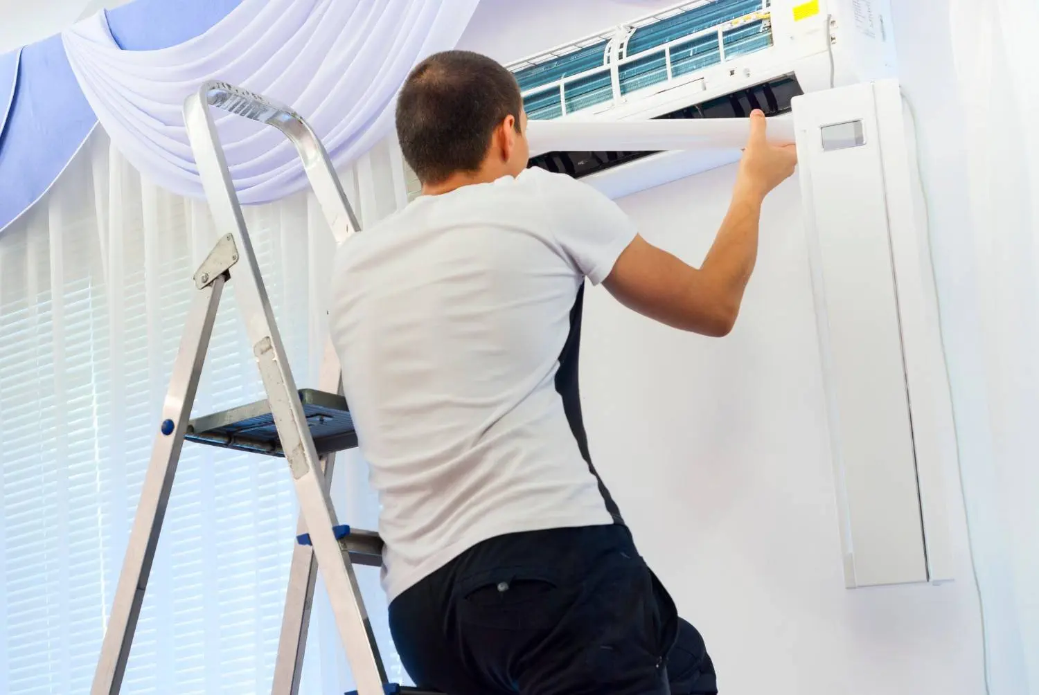 A person wearing a white t-shirt and dark pants is standing on a step ladder to service a white ductless mini-split indoor unit mounted high on a wall. They are handling a long, white plastic piece, likely part of the casing or air guide, while the unit's filters and internal components are visible. The background includes a window with white blinds and blue draped curtains.