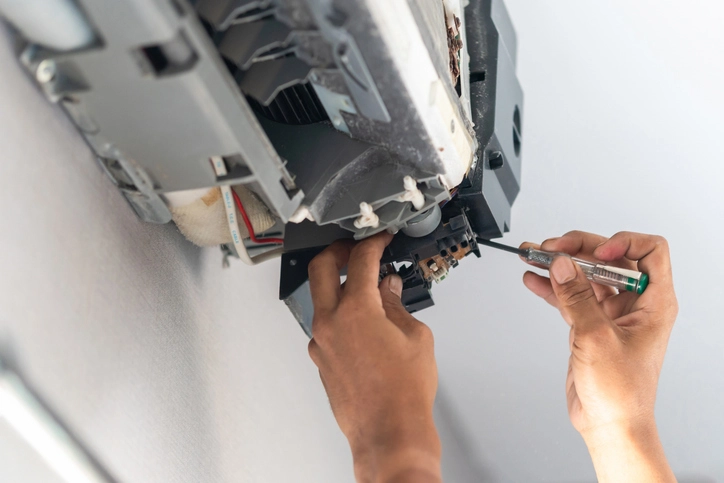 A close-up, low-angle shot shows a technician's hands using a small screwdriver to work on the exposed electrical components and wiring inside a disassembled, ceiling-mounted or high-wall ductless mini-split air conditioner indoor unit. The gray plastic casing and internal motor/fan assembly are visible.