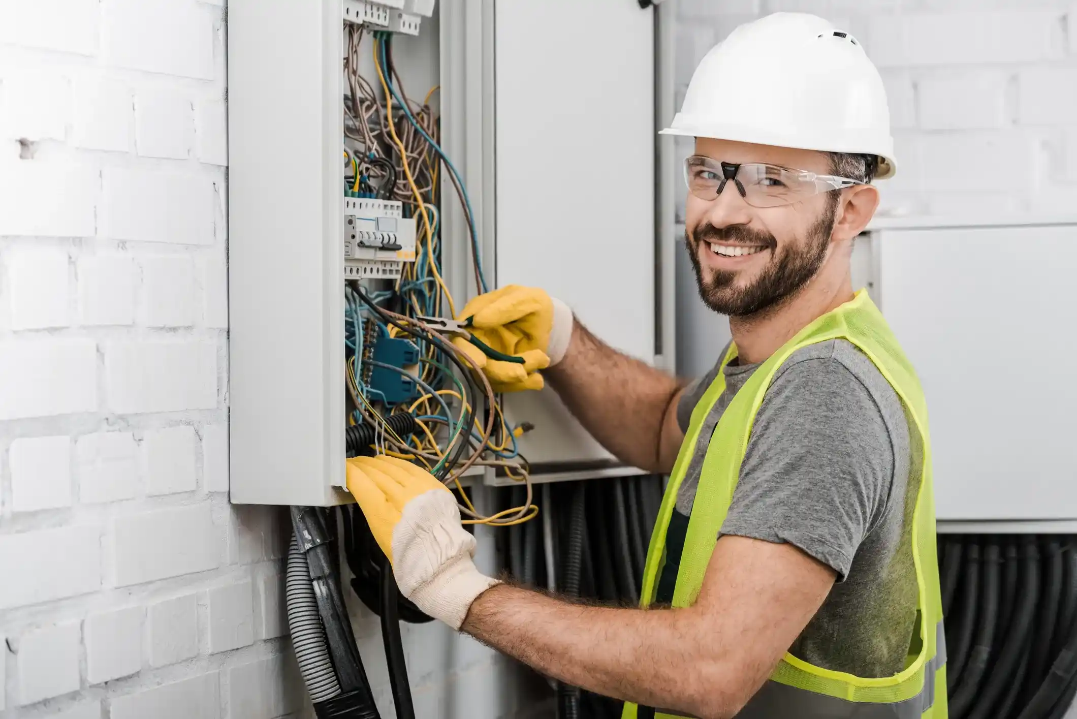 A smiling electrician in a white hard hat and safety vest works on a complex junction box with many wires.