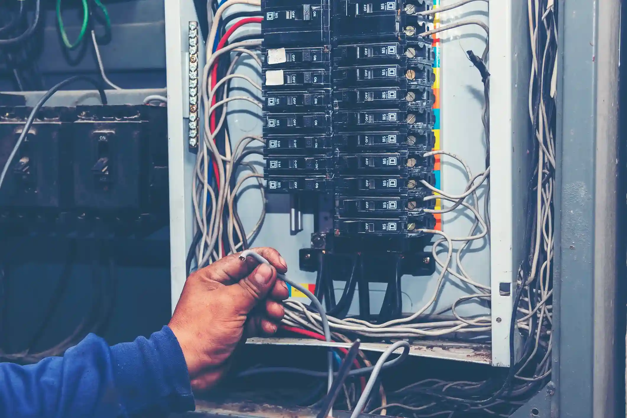 A close-up of a hand holding a stripped wire near a residential circuit breaker panel, showing rows of black breakers and a tangle of loose wires.