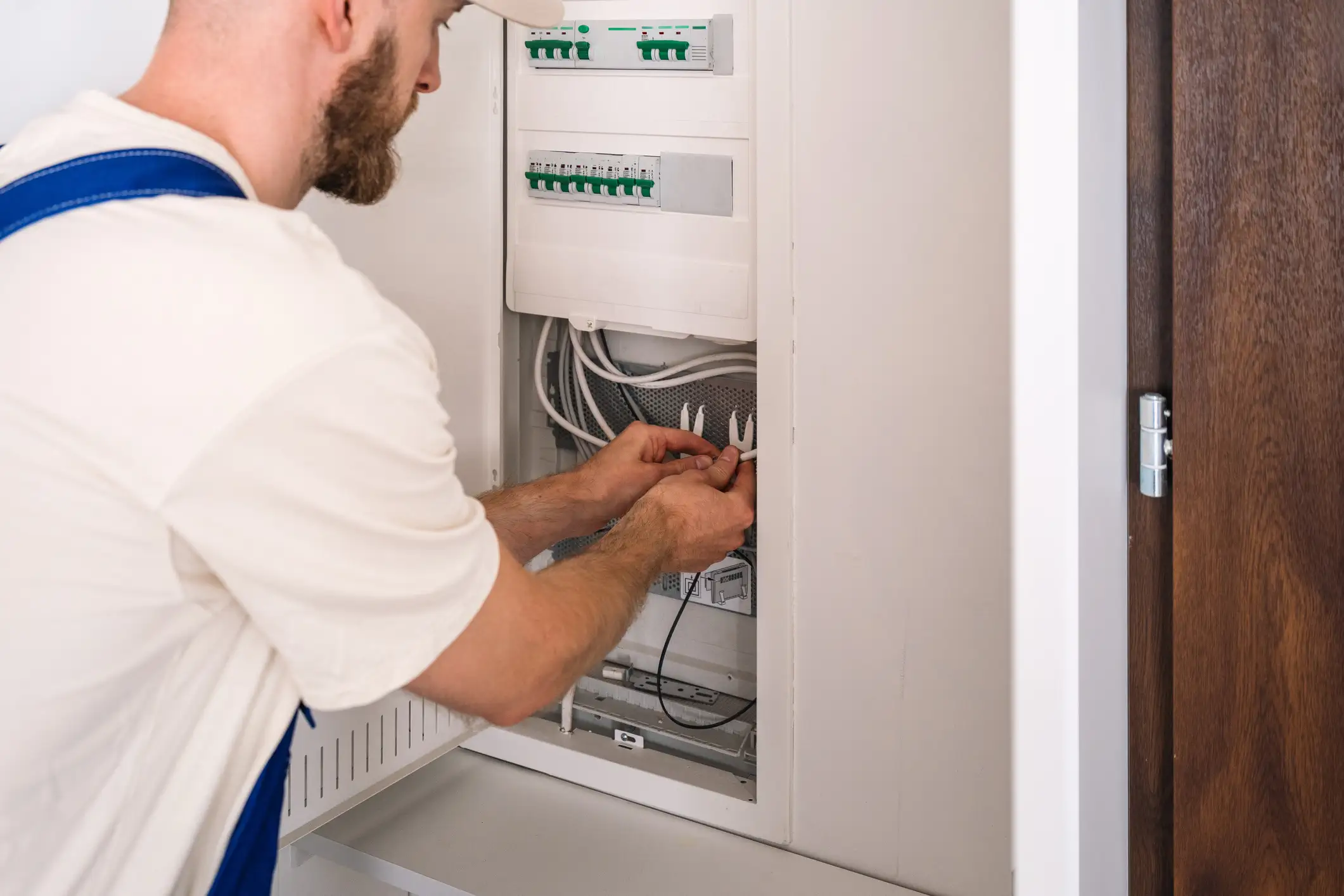 A male electrician with a beard, wearing white overalls, works on the wiring inside a modern residential electrical distribution board. His hands are connecting white cables near the bottom of the panel, with multiple green-switch circuit breakers visible above.