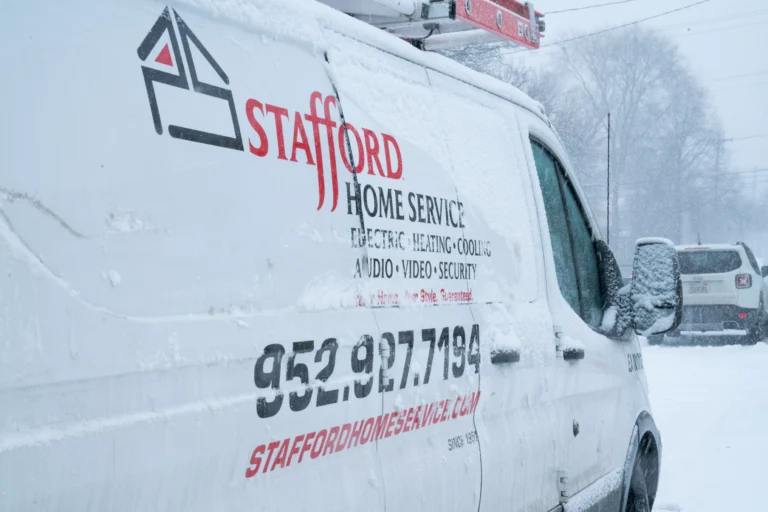 A close-up of a white service van, heavily covered in snow and ice, identifies it as belonging to Stafford Heating, Cooling, Plumbing & Electrical. The company logo, phone number (952.927.7194), and website (STAFFORDHOMESERVICE.COM) are visible on the driver's side door. The listed services include ELECTRIC, HEATING, COOLING, AUDIO, VIDEO, SECURITY. The background shows a heavily snow-covered residential street, suggesting the vehicle is working during severe winter weather. The image conveys dedication to providing home services regardless of the conditions.