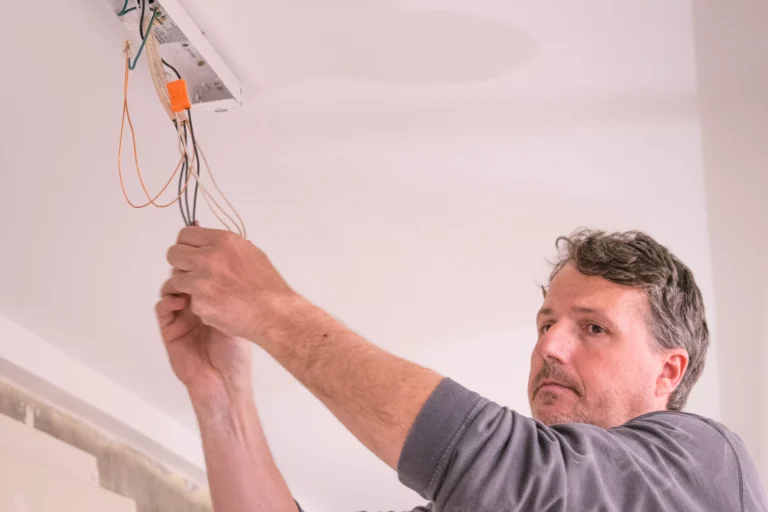 A close-up image shows a man, seen from the chest up, working on an electrical fixture mounted on a white ceiling. He has short, curly dark hair and is wearing a dark grey long-sleeved shirt. His hands are up, holding and manipulating a bundle of wires (black, white, and orange) that hang down from the light fixture housing. An orange wire nut/connector is visible attached to one set of wires inside the fixture.