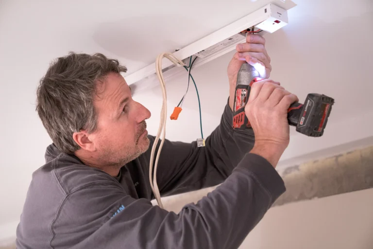 A man with salt-and-pepper hair, wearing a dark grey shirt, is installing or repairing a light fixture on a white ceiling. He is using a cordless drill/driver with a built-in light to secure the fixture housing to the ceiling. Several wires, including a bundle connected with an orange wire nut, are visible hanging down next to the fixture.