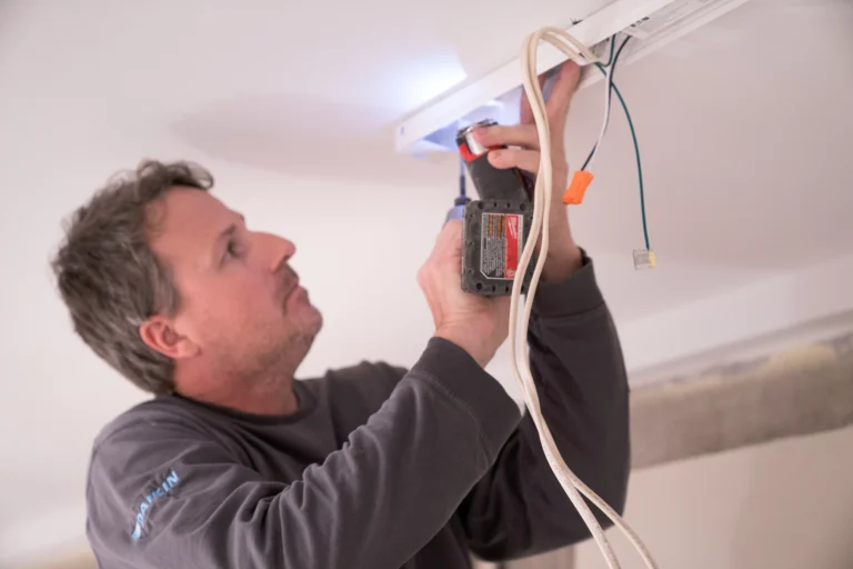 A man with medium-length grey hair and a light beard, wearing a dark grey long-sleeved shirt, is installing or adjusting a white light fixture mounted on a white ceiling. He is using a cordless drill/driver, which is illuminated by its built-in light, to fasten the fixture. Several wires, including a white bundled cable and a loose green wire with an orange wire nut, are visible hanging near the fixture. The sleeve of his shirt has a light blue logo or writing, possibly "DAIKIN."
