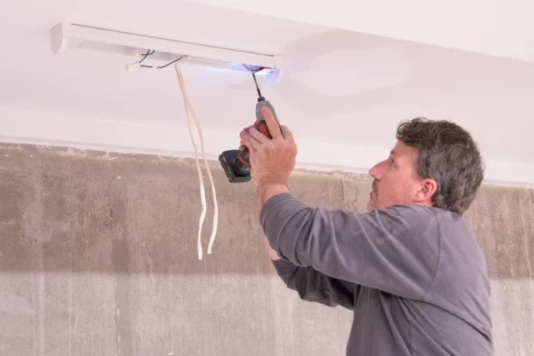 A man wearing a dark grey long-sleeved shirt is installing a white linear light fixture onto a white ceiling. He is using a cordless drill/driver to secure the fixture, with the tool's light illuminating the area. Several electrical wires are hanging down from the ceiling near the fixture. The wall below the ceiling is unfinished and features a rough, grey concrete texture.
