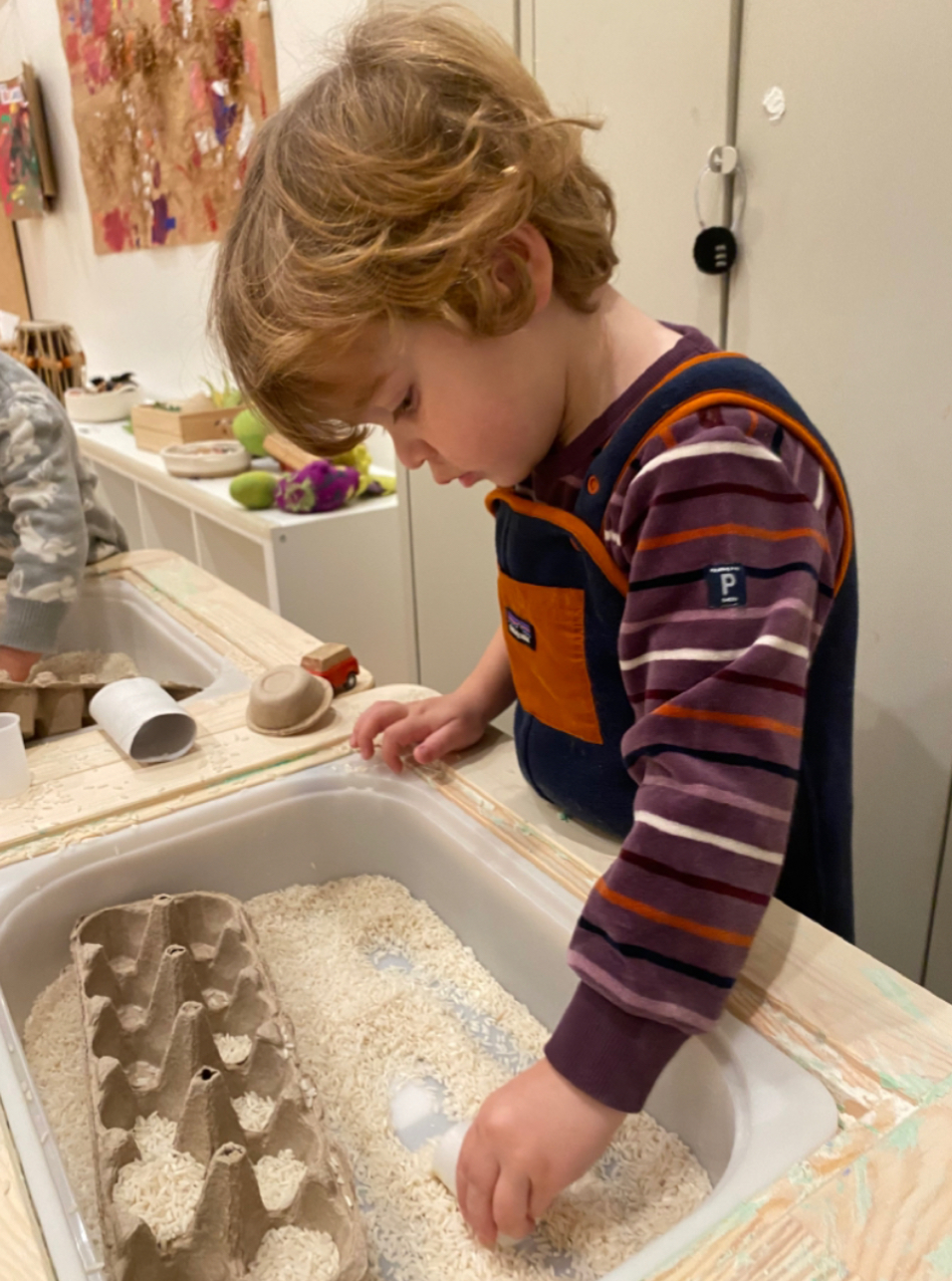 Young child playing with a sensory bin filled with rice and egg carton pieces indoors.