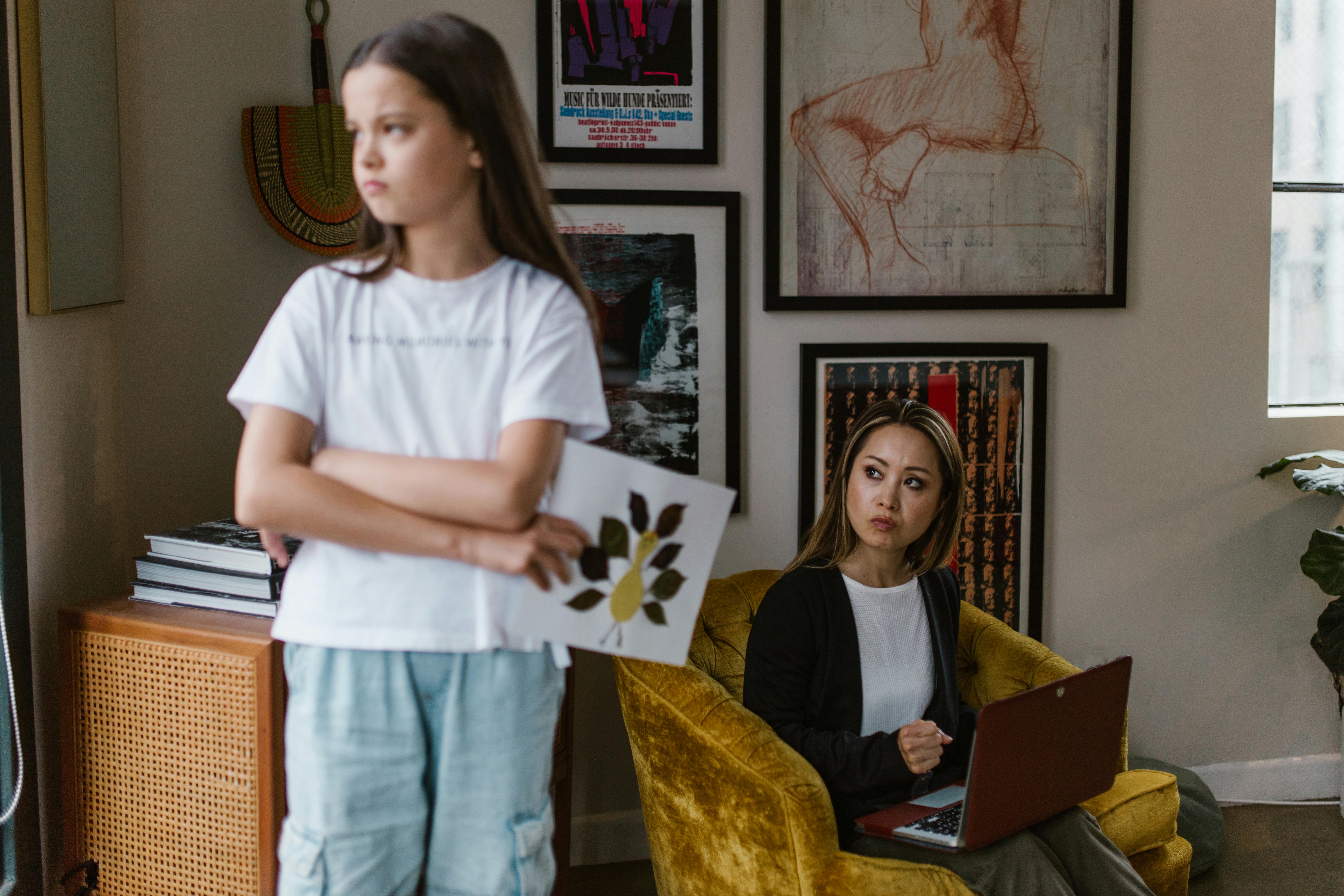 Young girl with crossed arms holding a drawing turned away from a seated woman with a laptop.