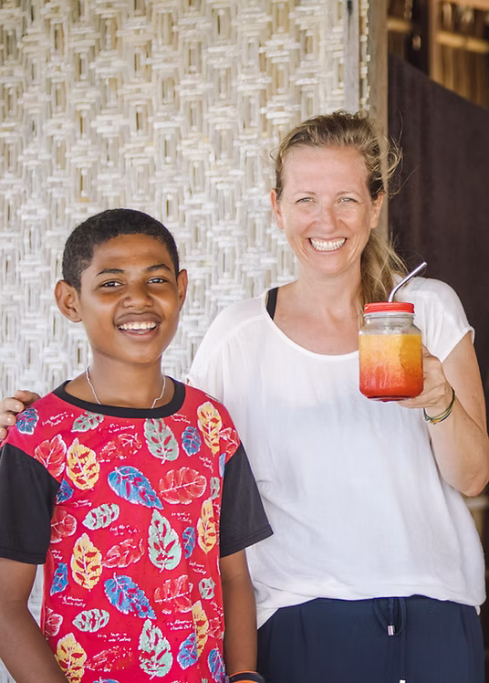 Lächelnde Frau hält ein Glas mit einem bunten Getränk, steht neben einem lächelnden Jungen in einem roten T-Shirt mit buntem Blättermuster.