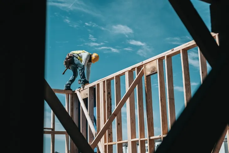 Construction worker in yellow hard hat building wooden frame against blue sky
