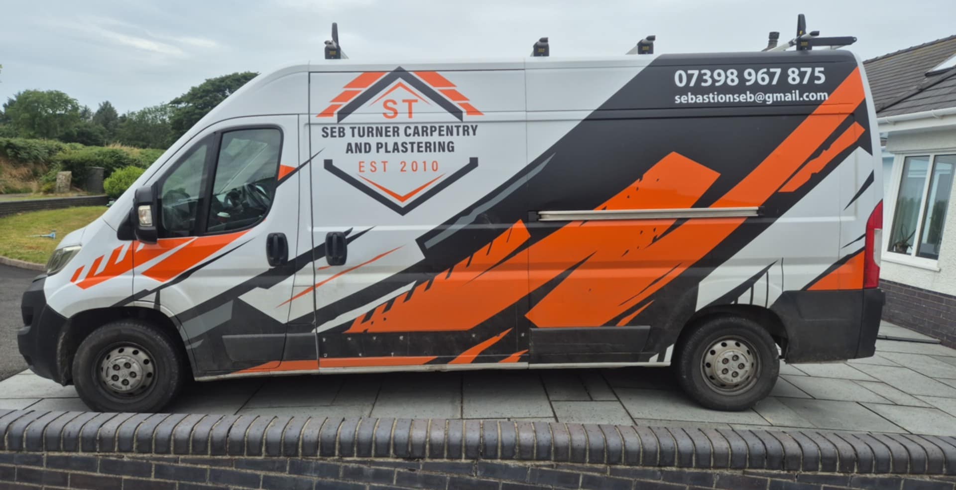 White work van with orange and black geometric graphics and the logo for Seb Turner Carpentry and Plastering on the side. The van displays the business name, contact phone number, email address, and 'EST 2010'. The van is parked on a paved driveway next to a house.