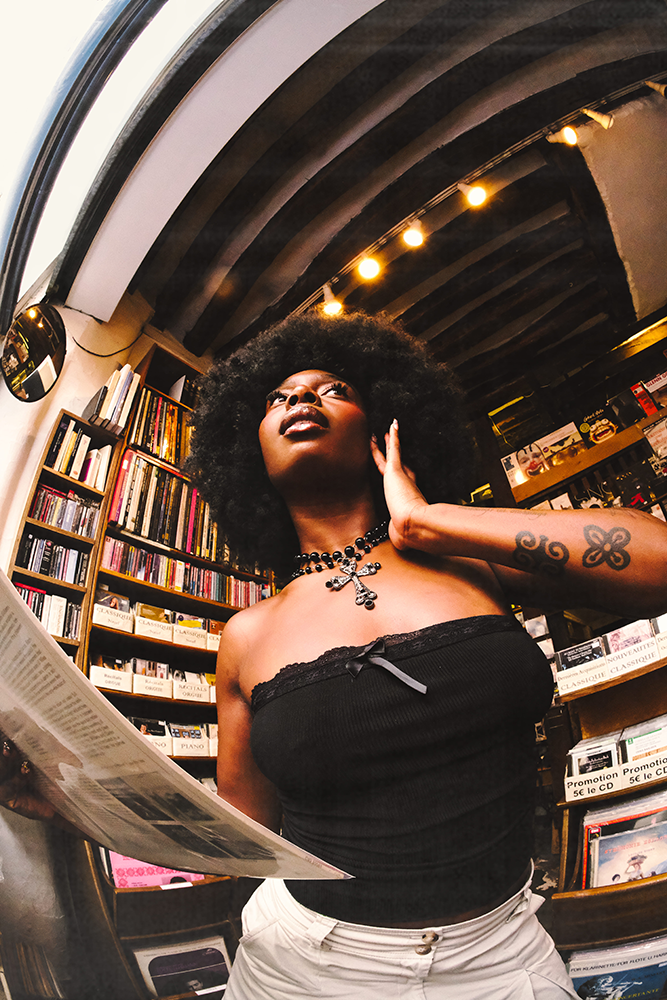 Photographie de Romane Kouo. Portrait d'une femme dans une librairie avec un journal à la main. 