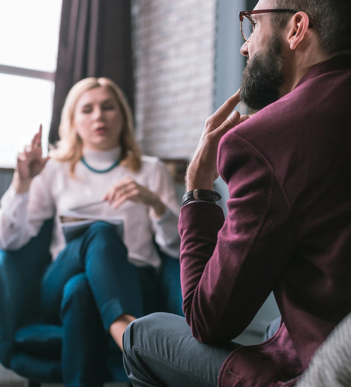 Man with beard and glasses sitting thoughtfully while talking to a woman taking notes in a cozy room.