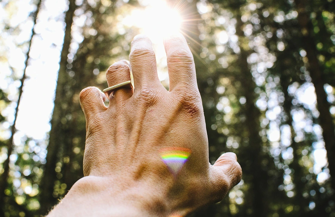 Hand with a ring reaching towards the sun in a forest with a small rainbow reflection on the skin.