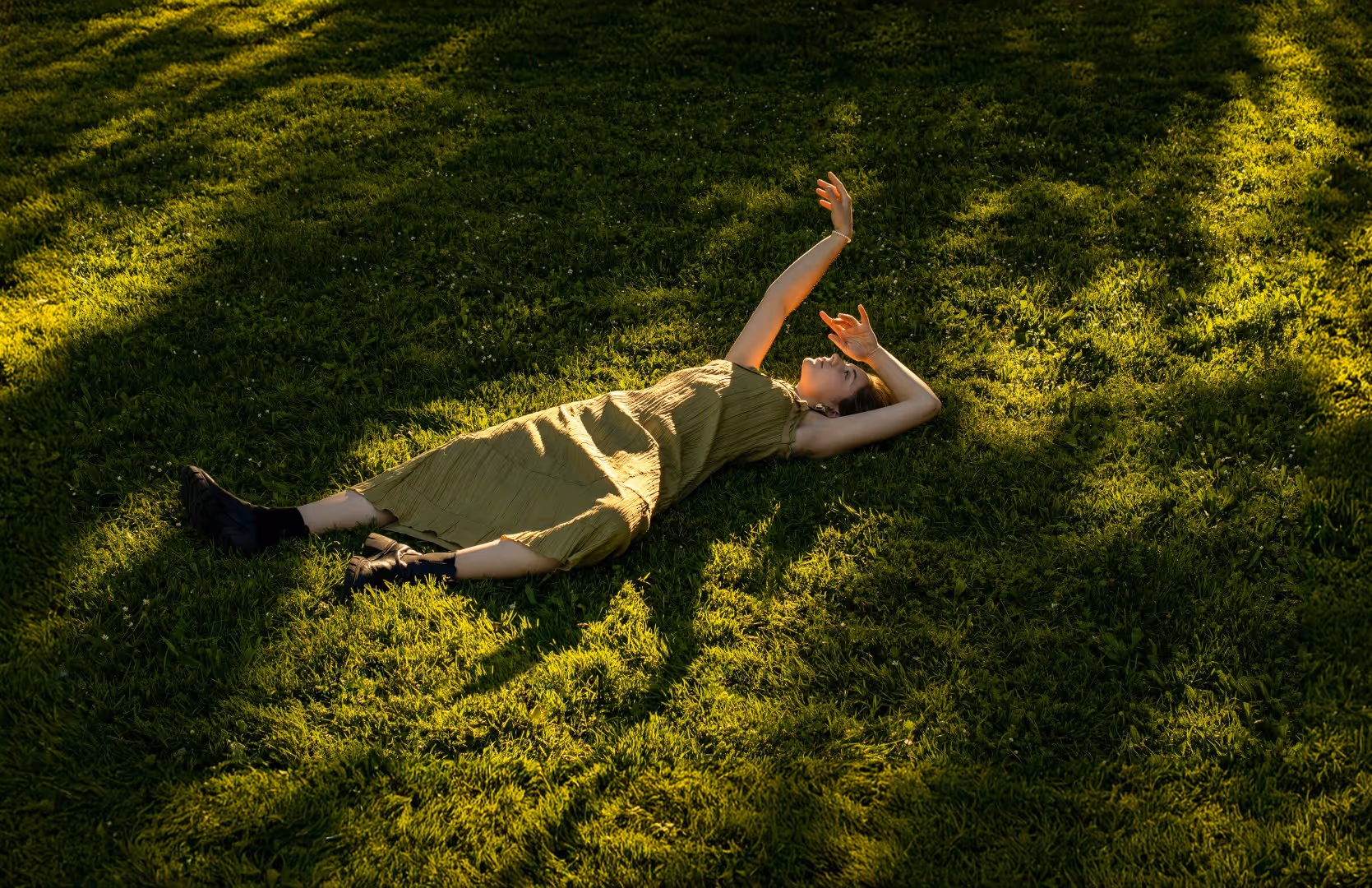Woman in green dress lying on grass with arms raised, in sunlight and shadows.