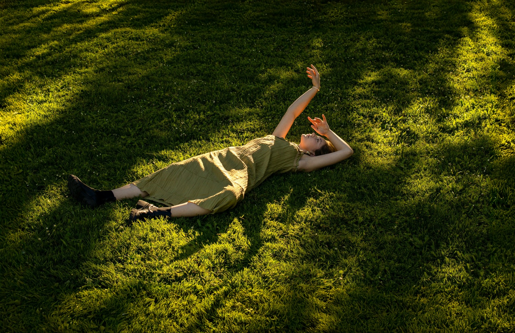 Woman in green dress lying on grass with arms raised, in sunlight and shadows.