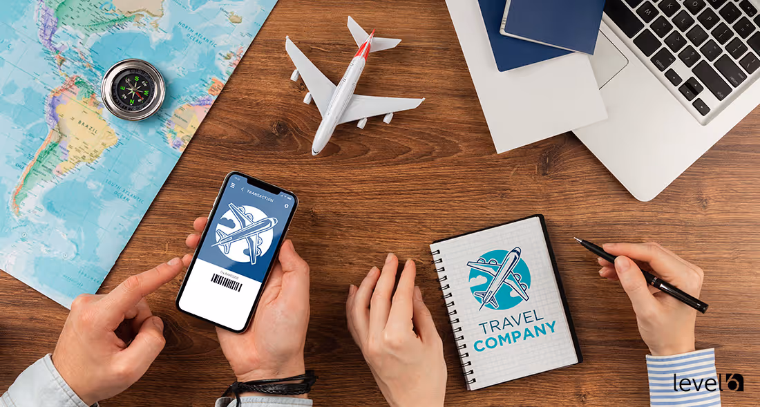 Hands holding a smartphone with a flight ticket app, a toy airplane, compass on a world map, a notebook with travel company logo, and a laptop on a wooden desk.
