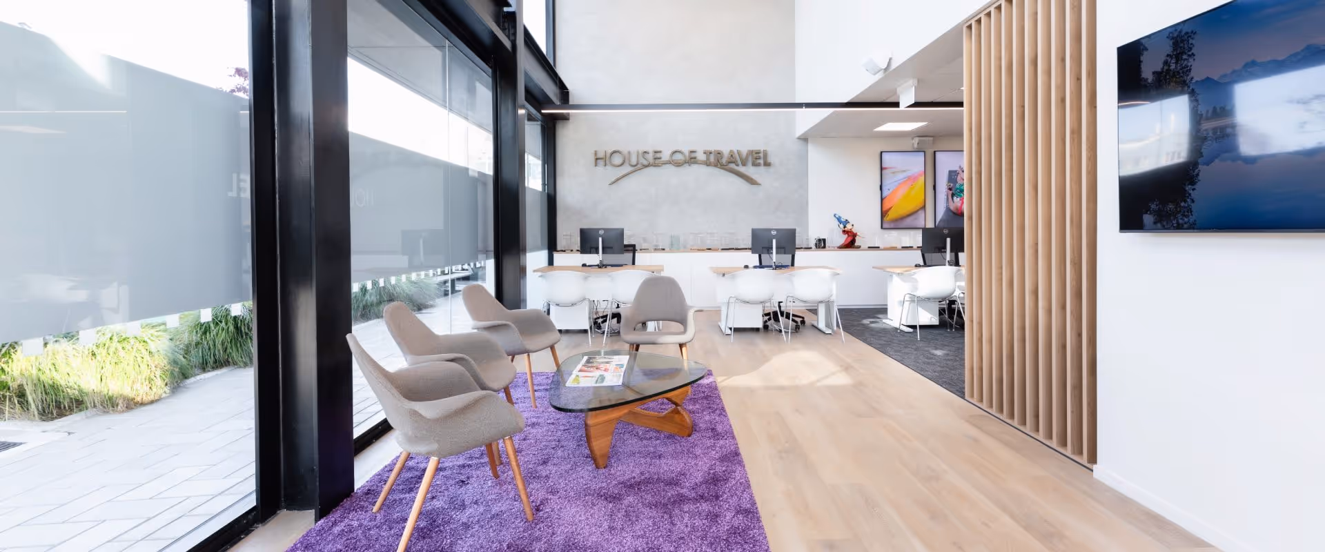 Modern travel agency office with a purple carpet seating area, glass coffee table, and workstations against the back wall under a House of Travel sign.