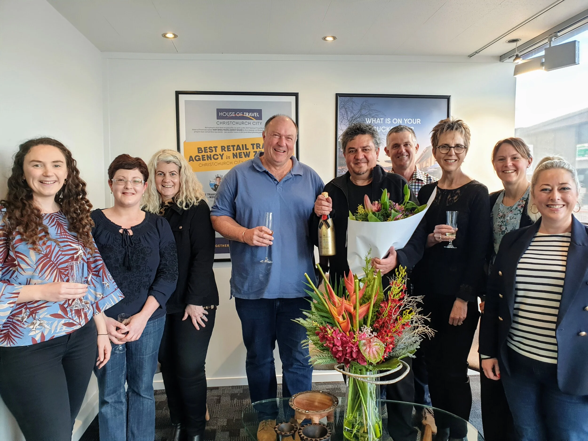 Group of nine people smiling indoors holding champagne glasses, with one person holding a bottle and another holding a bouquet of flowers, standing behind a glass table with a large floral arrangement.