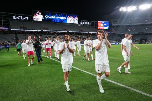 Soccer players in white uniforms and 'Qualified' shirts clapping and walking on the field in a stadium at night.