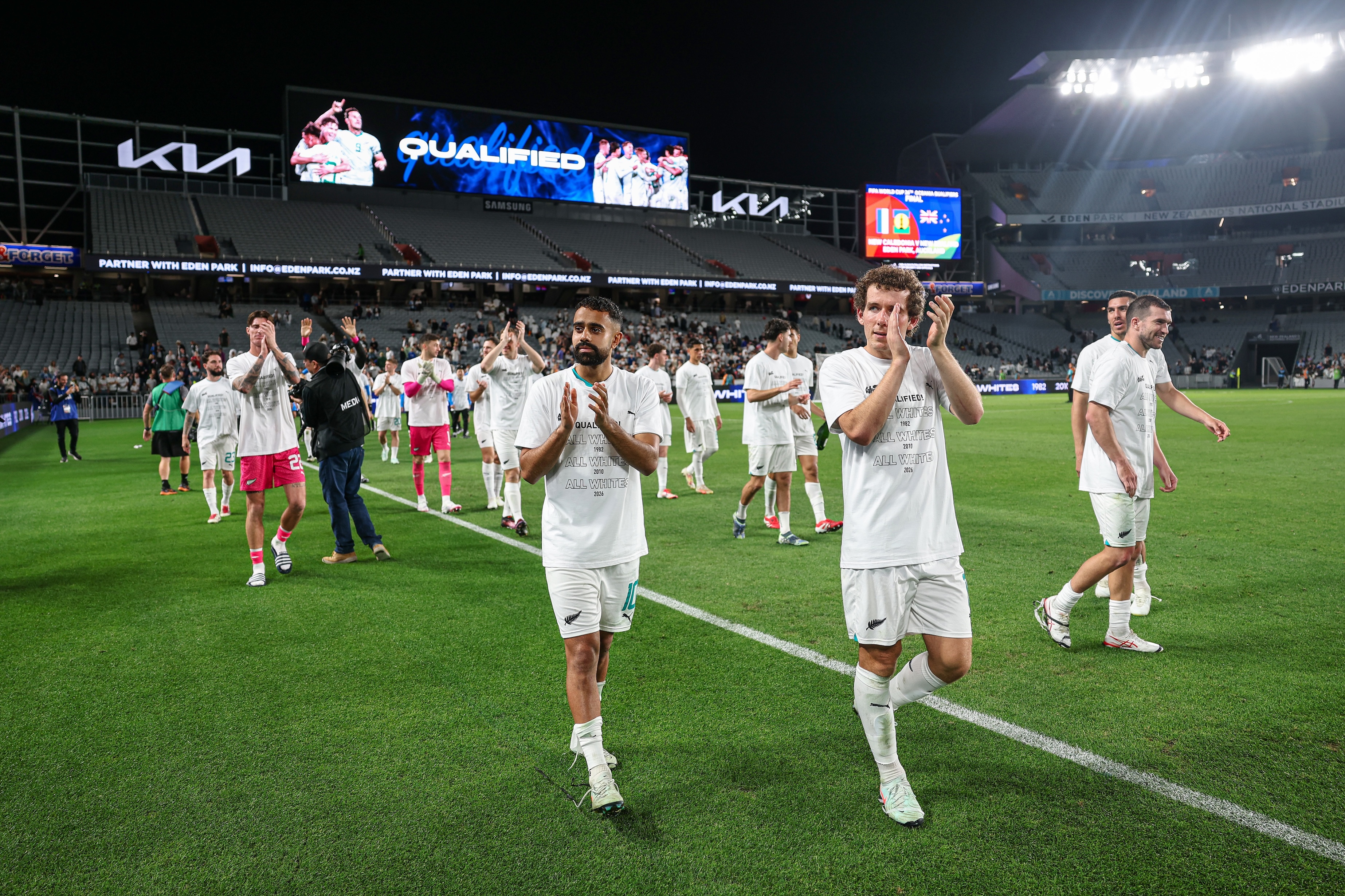 Soccer players in white uniforms and 'Qualified' shirts clapping and walking on the field in a stadium at night.