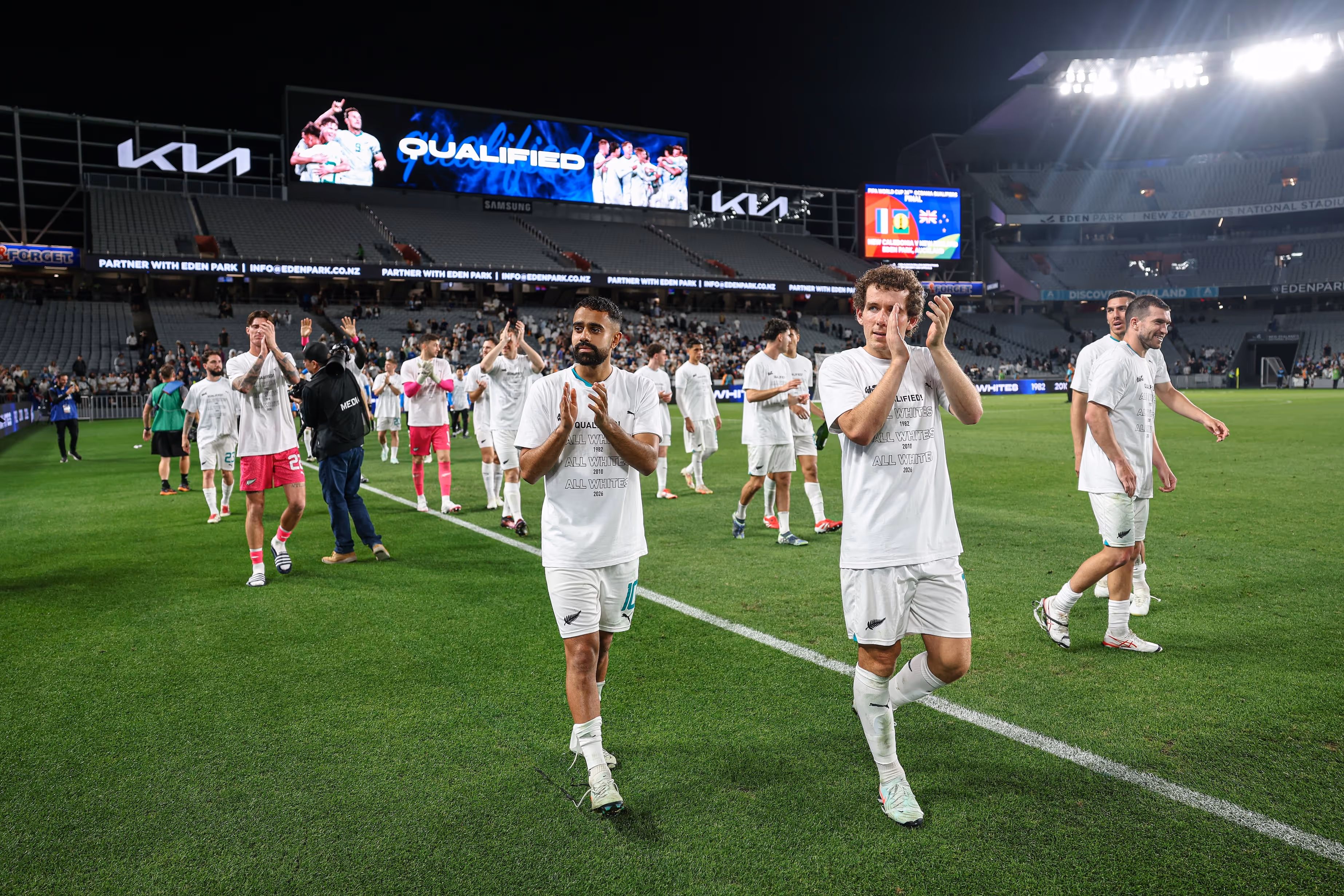 Soccer players in white uniforms and 'Qualified' shirts clapping and walking on the field in a stadium at night.