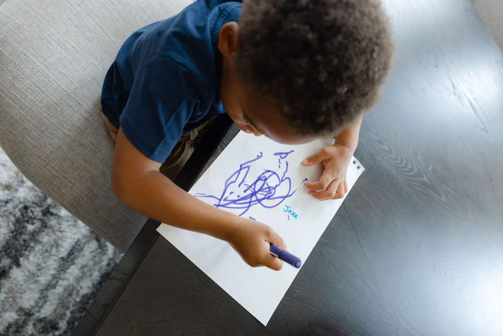 A child leaning over a desk, writing on a blank piece of paper with a coloring pen.