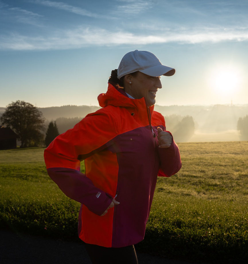 Doris läuft Bajoli bei aufgehender Sonne vor Wiesen und Bäumen im Morgentau.