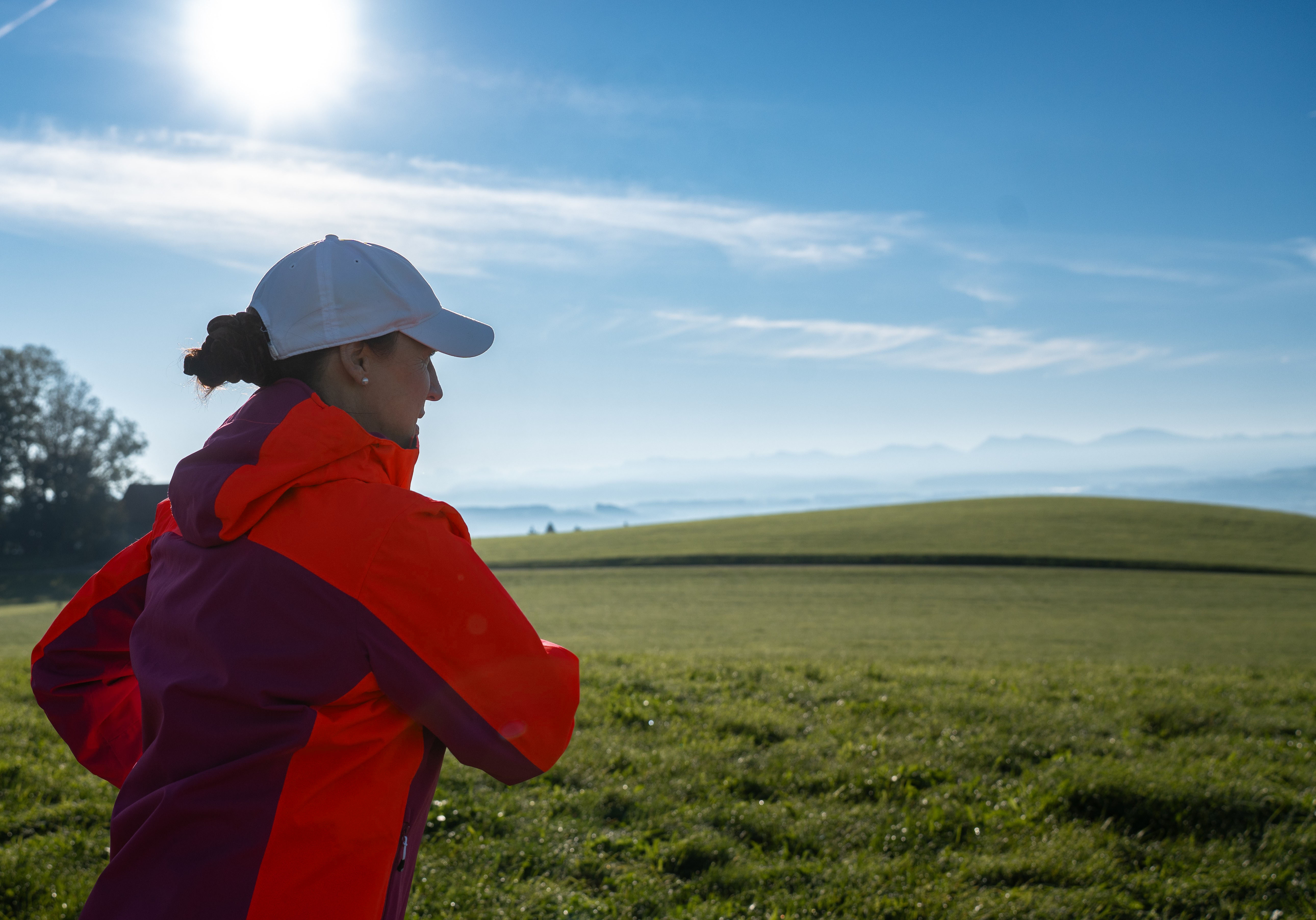 Doris von hinten beim Bajoli-Lauf. Vor ihr grüne Wiese und eine Bergkulisse bei blauem Himmel und Sonnenschein.